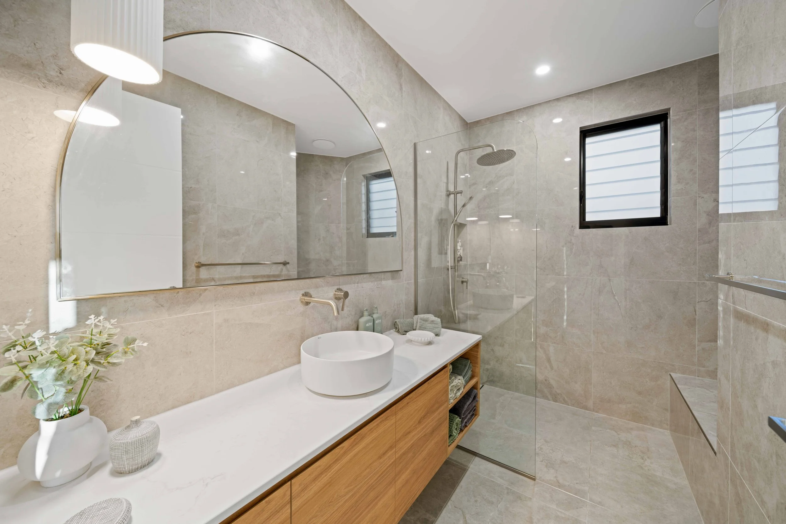 Modern bathroom with beige tiled walls and floor, large mirror, wooden vanity with white countertop, round vessel sink, and glass-enclosed shower with rainfall showerhead and small window.