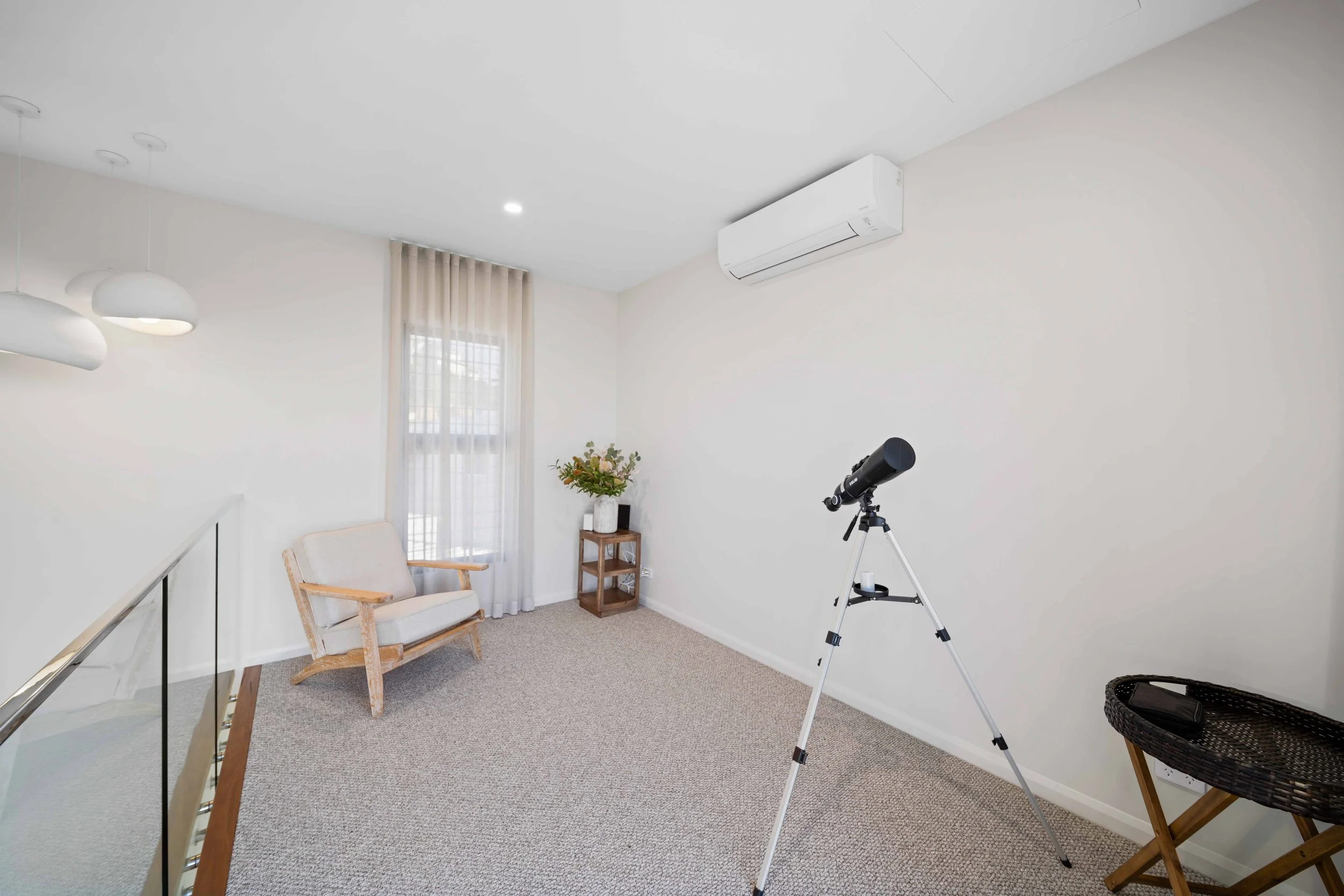 Minimalist room with white walls, beige carpet, a telescope on a tripod, a small wooden side table with a bouquet of flowers, a beige armchair near a window with sheer curtains, and a black basket on a wooden stand.