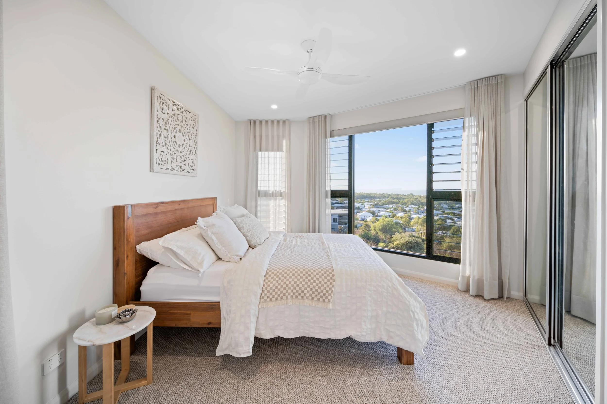 A bedroom with a large window overlooking a cityscape, featuring a wooden bed with white bedding and multiple pillows, a small marble side table with candles, and a mirrored closet door.