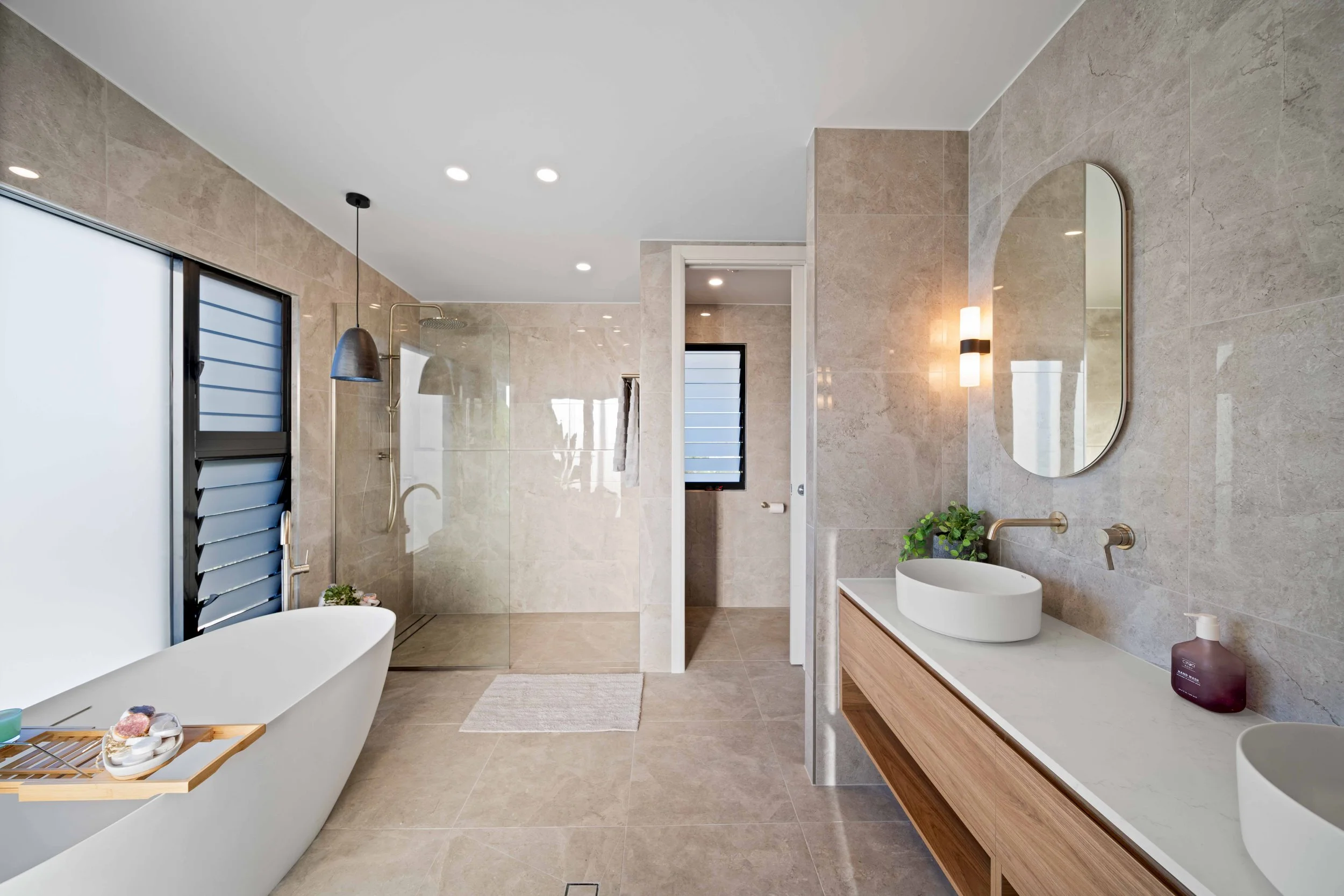 Modern bathroom with a freestanding bathtub, walk-in shower with glass partition, large mirror, wooden vanity with vessel sinks, and neutral-toned tiles on walls and floor.
