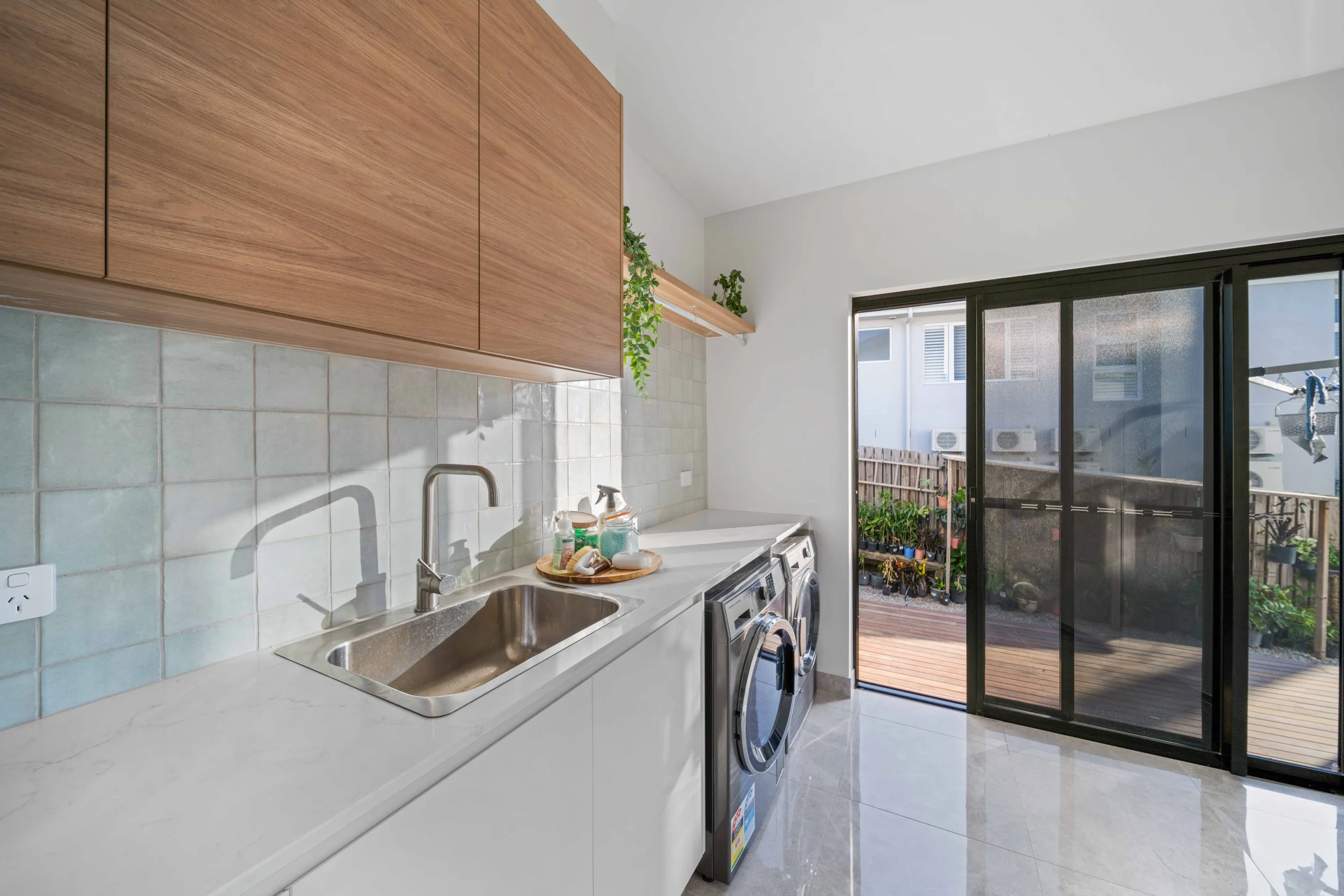 A laundry room with a white countertop, a stainless steel sink, wooden cabinets, a washing machine, a dryer, a small shelf with potted plants, and a large sliding glass door opening to a small outdoor deck with potted plants.
