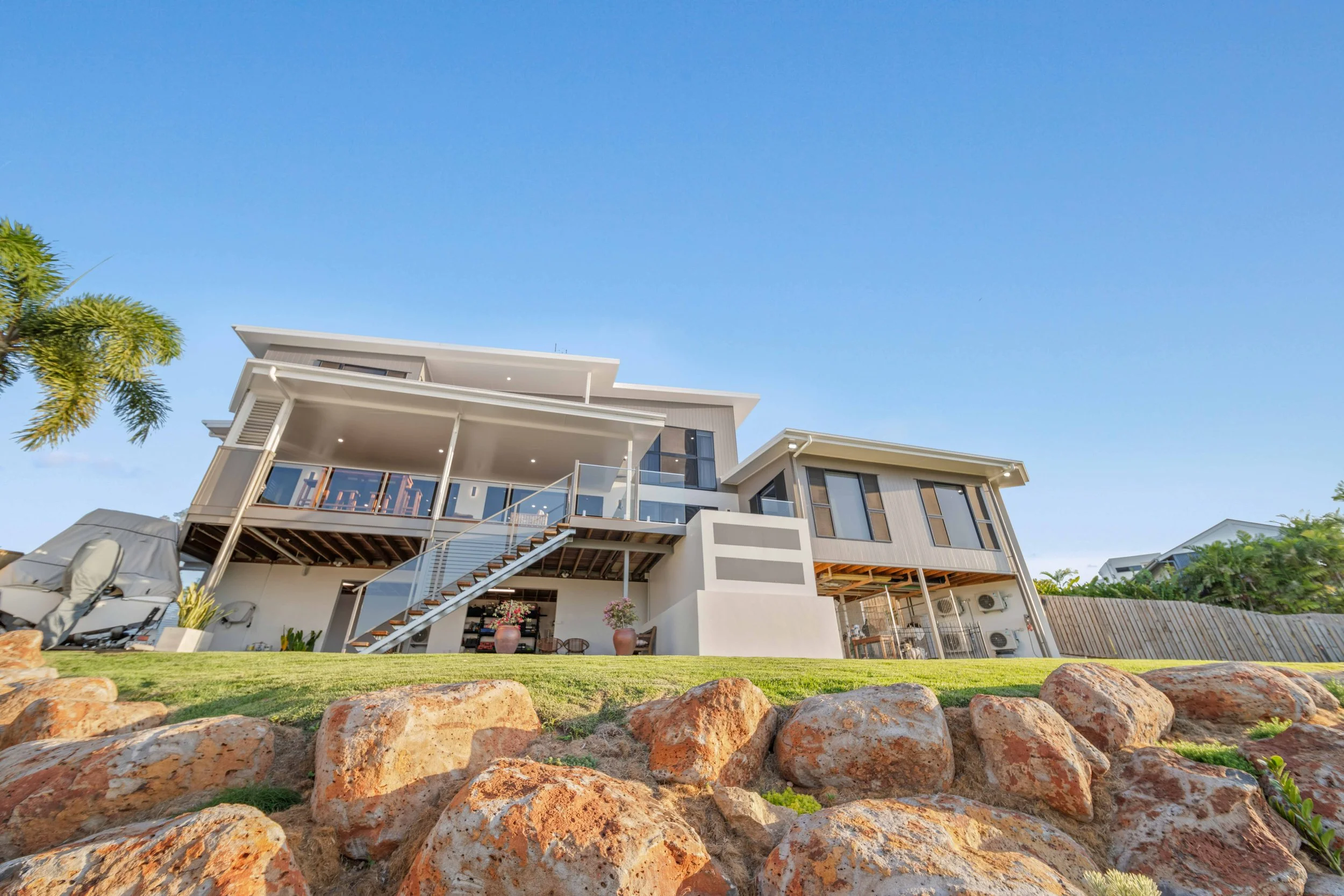 Modern multi-story house with large glass balconies, set against a clear blue sky, with rocky landscaping in the foreground and some trees on the side.