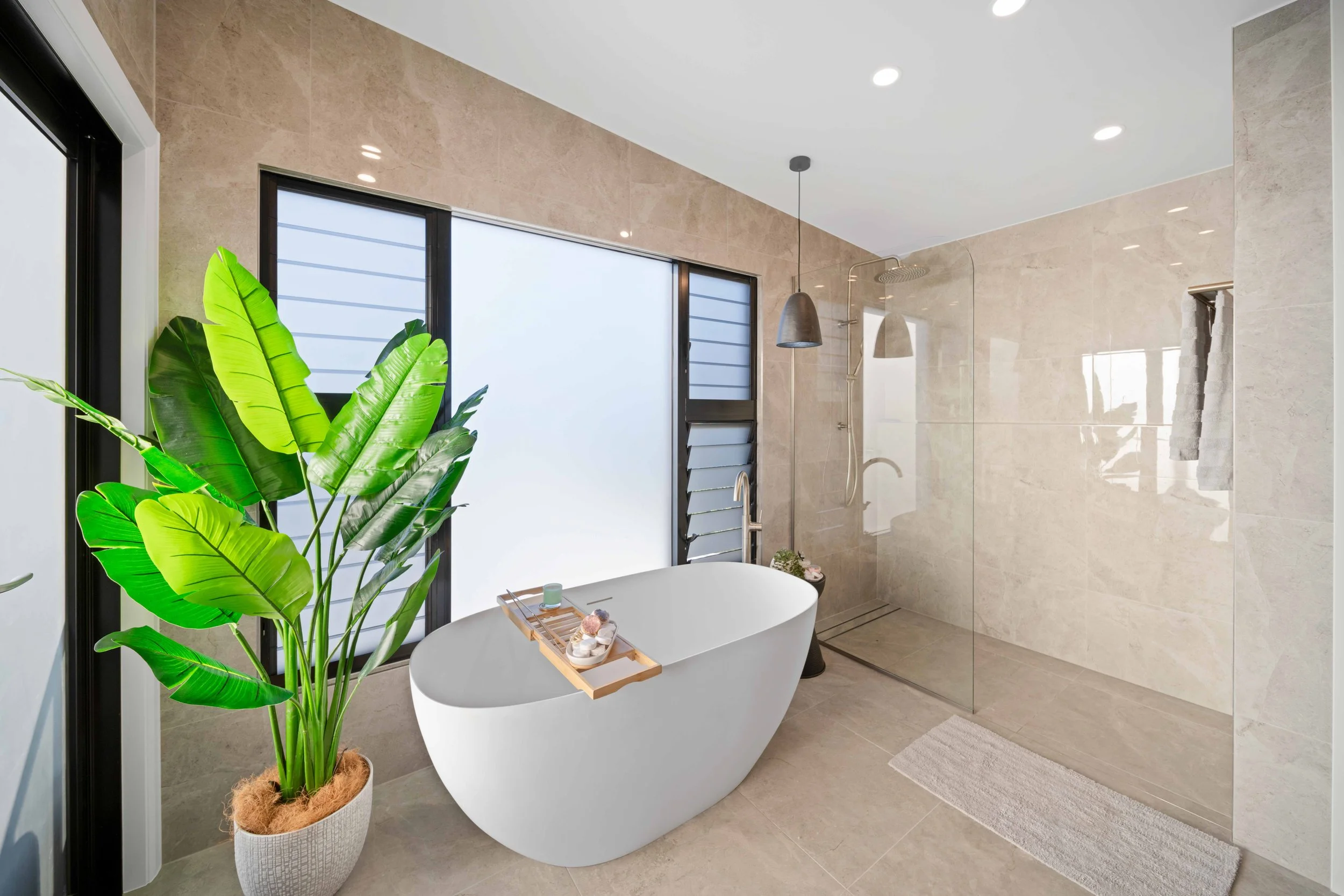 Modern bathroom with a white freestanding bathtub, large green potted plant, and a glass-enclosed shower area with beige tiled walls and floor. Features frosted windows, hanging pendant lights, and a white bath mat.