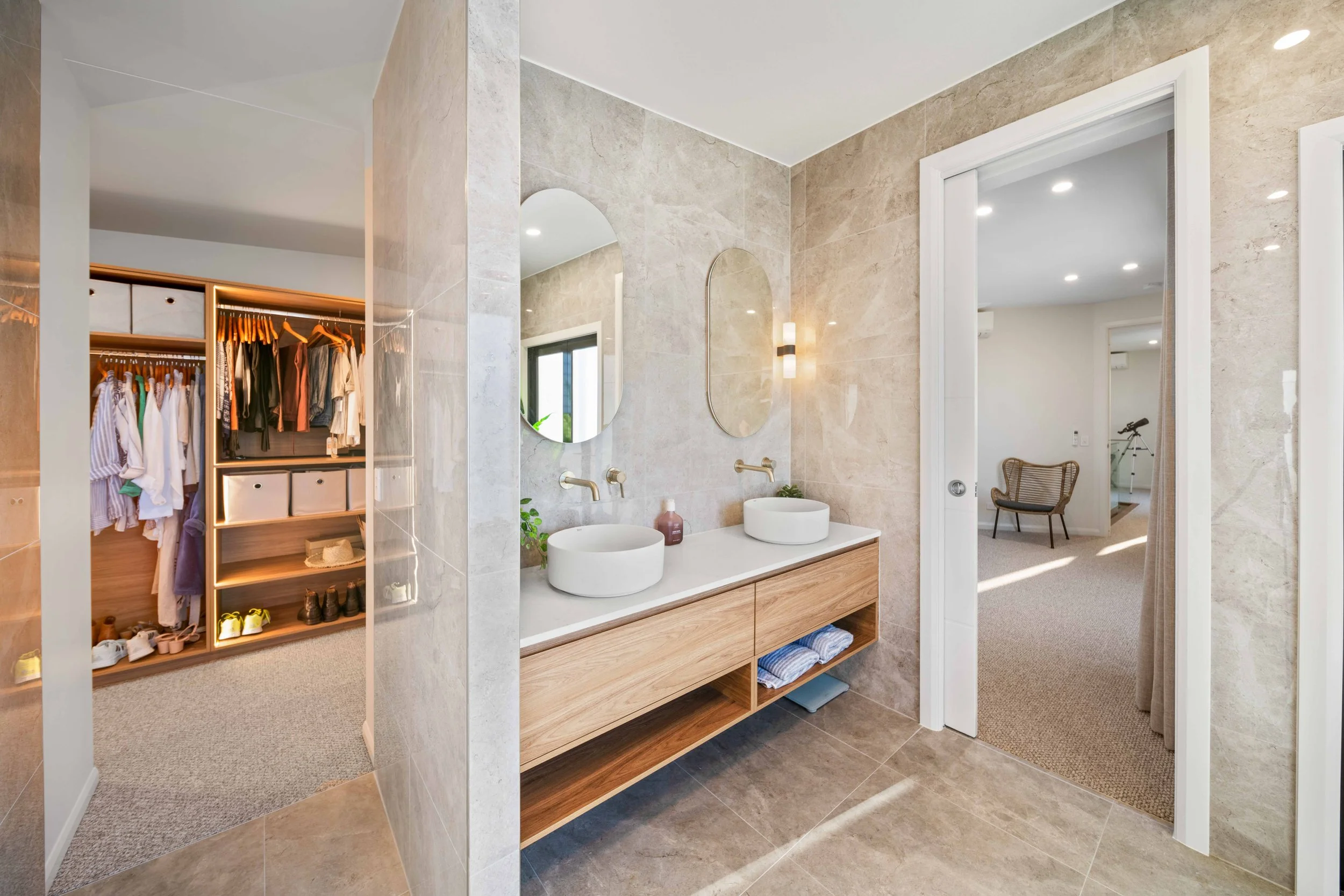 Modern bathroom with dual white vessel sinks, oval mirrors, beige tiled walls, and a view into a walk-in closet and a sitting area with a chair.