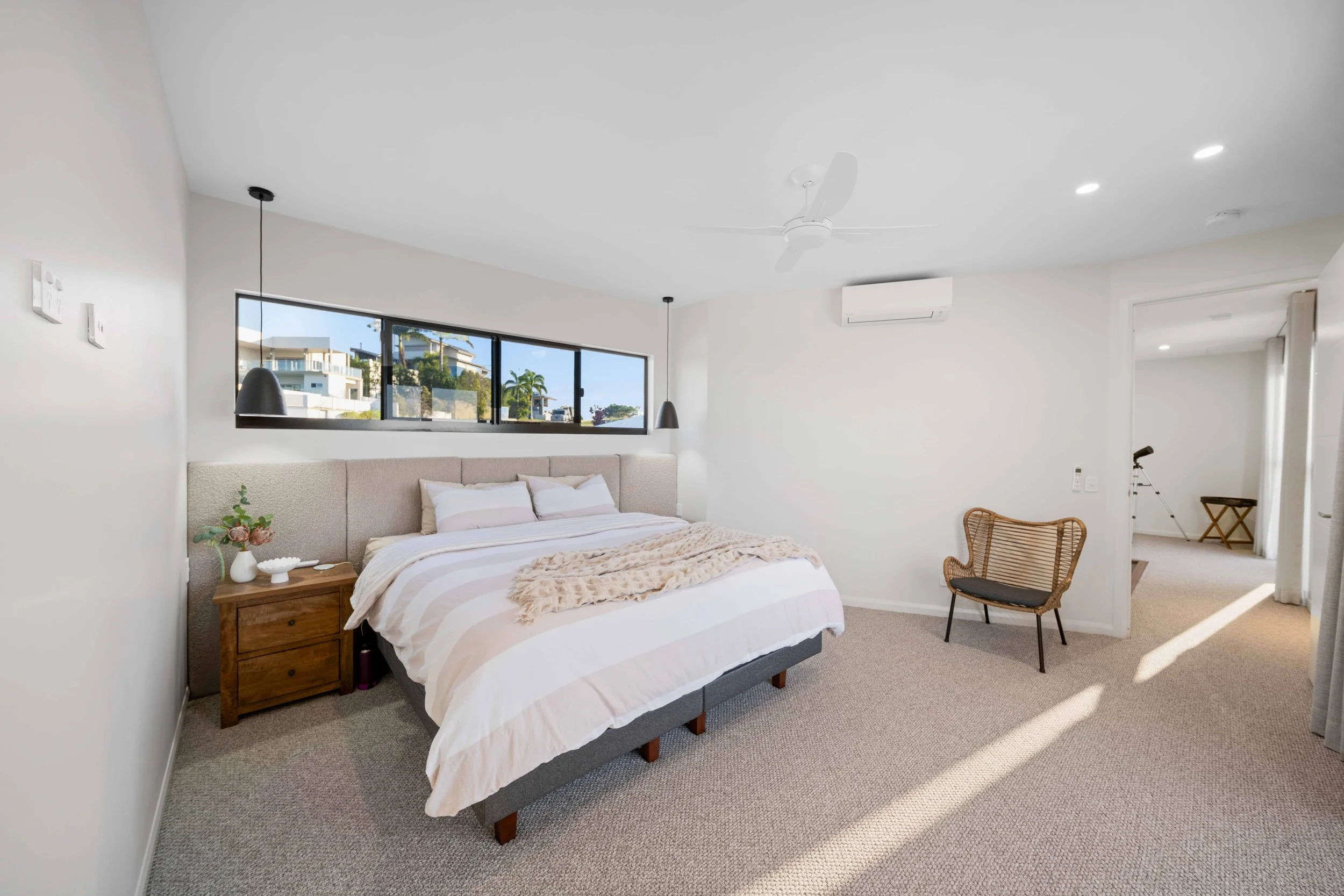 Bright bedroom with white walls, beige carpet, and a large window with outdoor view. Contains a bed with white bedding, a wooden nightstand with decorative items, two black pendant lights, and a wicker chair.