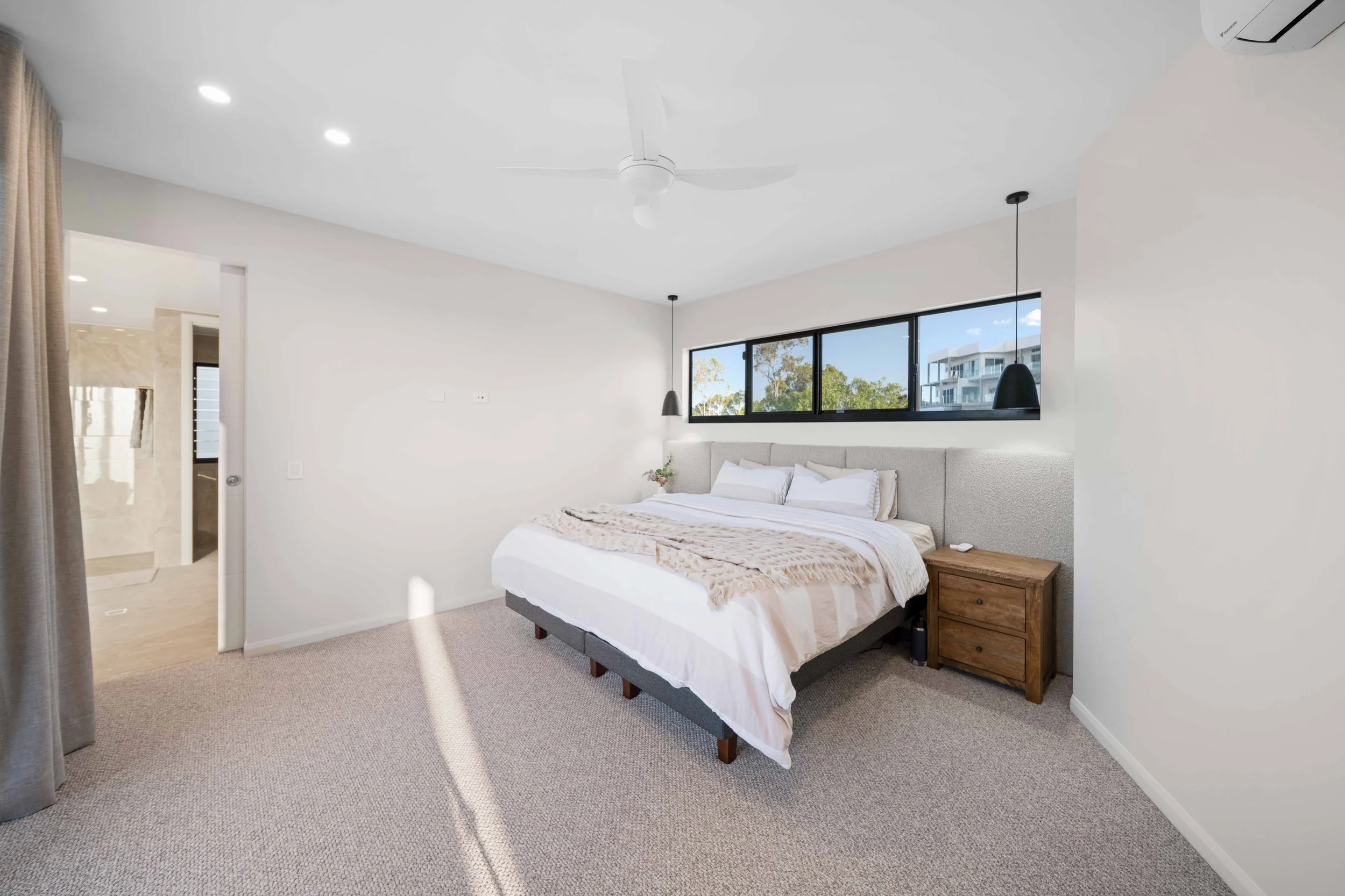 Modern bedroom with a large bed, white bedding, bedside table, black pendant lights, and a window showing trees and a building outside.