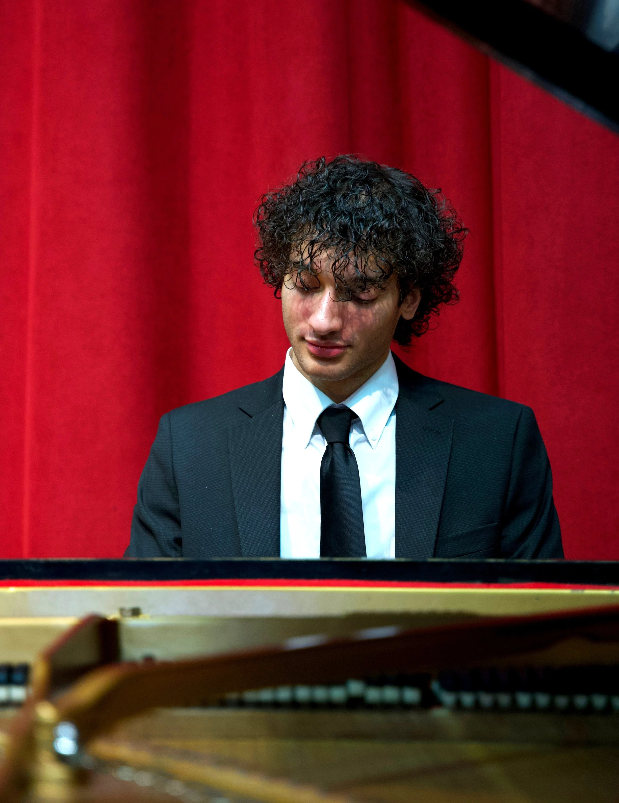 Oliver Rivera is playing a grand piano on stage with a red curtain backdrop.