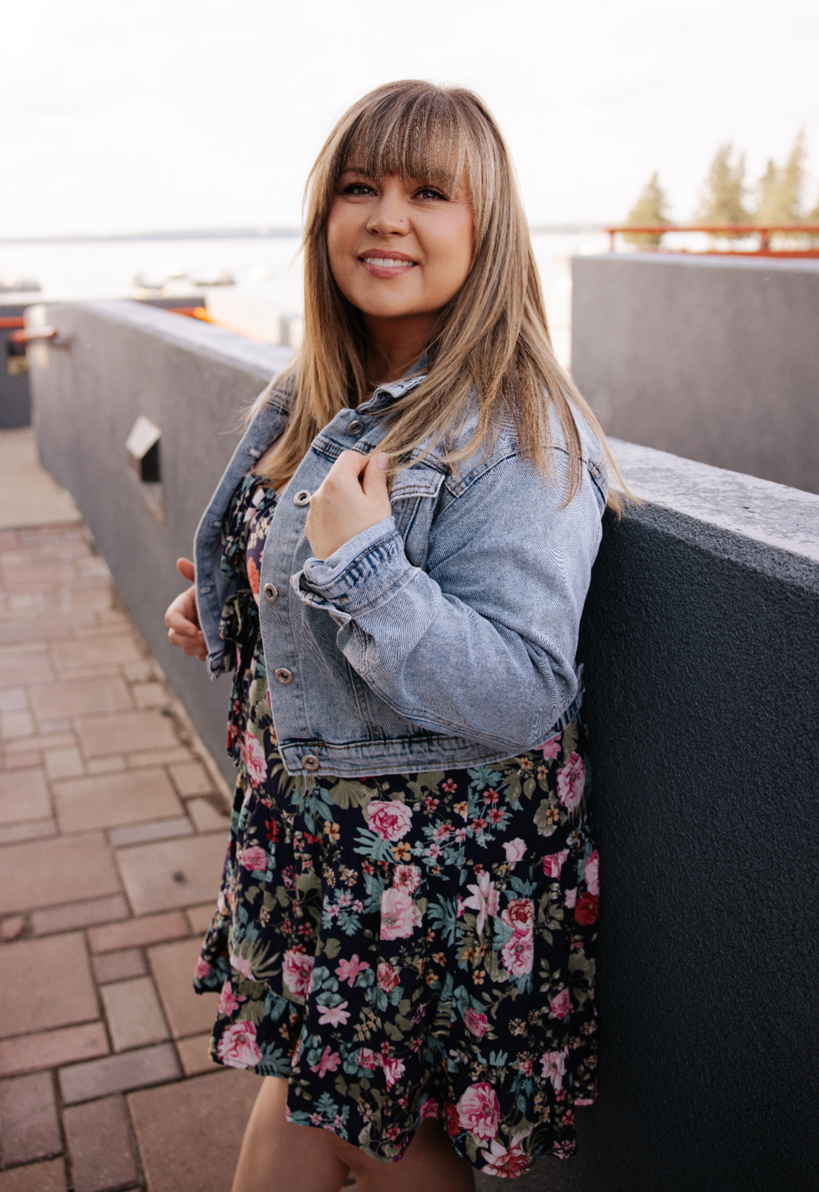 A woman with light brown hair wearing a denim jacket and floral dress, standing outdoors near a black wall with a view of water and boats in the background.