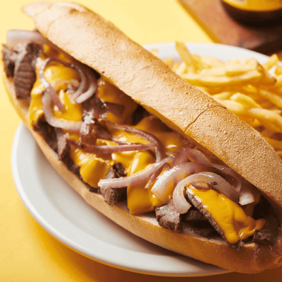Philly cheesesteak sandwich with grilled beef, melted cheese, and onions in a hoagie roll, served with French fries on a white plate.