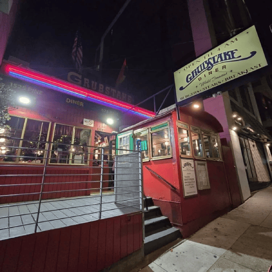 Nighttime view of a restaurant and bar with illuminated signs, including a large sign for 'Grubstake Restaurant' advertising burgers, steaks, and breakfast, and a neon Budweiser sign in a window, with parked cars and a dark sky in the background.