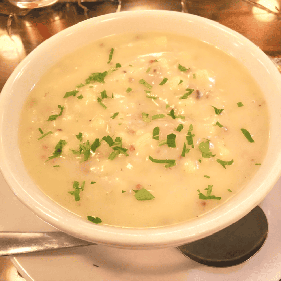 Bowl of creamy potato soup garnished with chopped parsley, served on a white plate with a metal spoon.