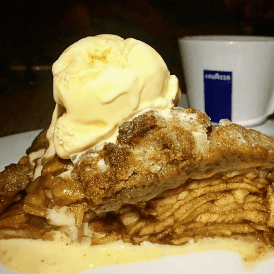 A slice of apple pie with a scoop of vanilla ice cream on top, served on a white plate with a cup of coffee in the background.