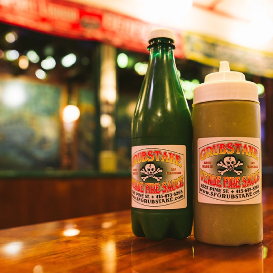 Two bottles of Grubstake Verde Fire Sauce on a polished wooden counter, featuring a vibrant label with skull logo and San Francisco branding, with a blurred restaurant background.