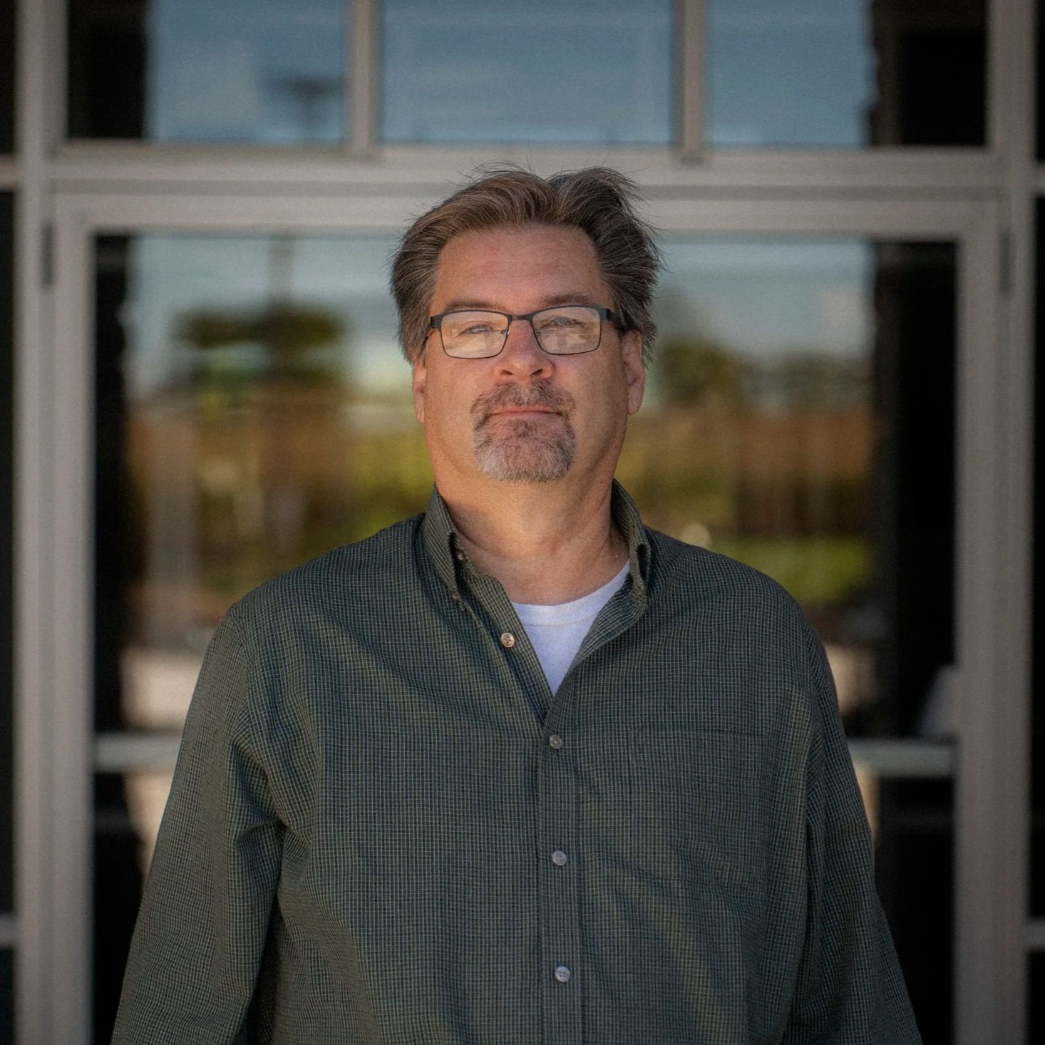 A middle-aged man with dark hair, glasses, and facial hair, wearing a striped button-down shirt with a small Ralph Lauren logo, standing against a light gray brick wall.