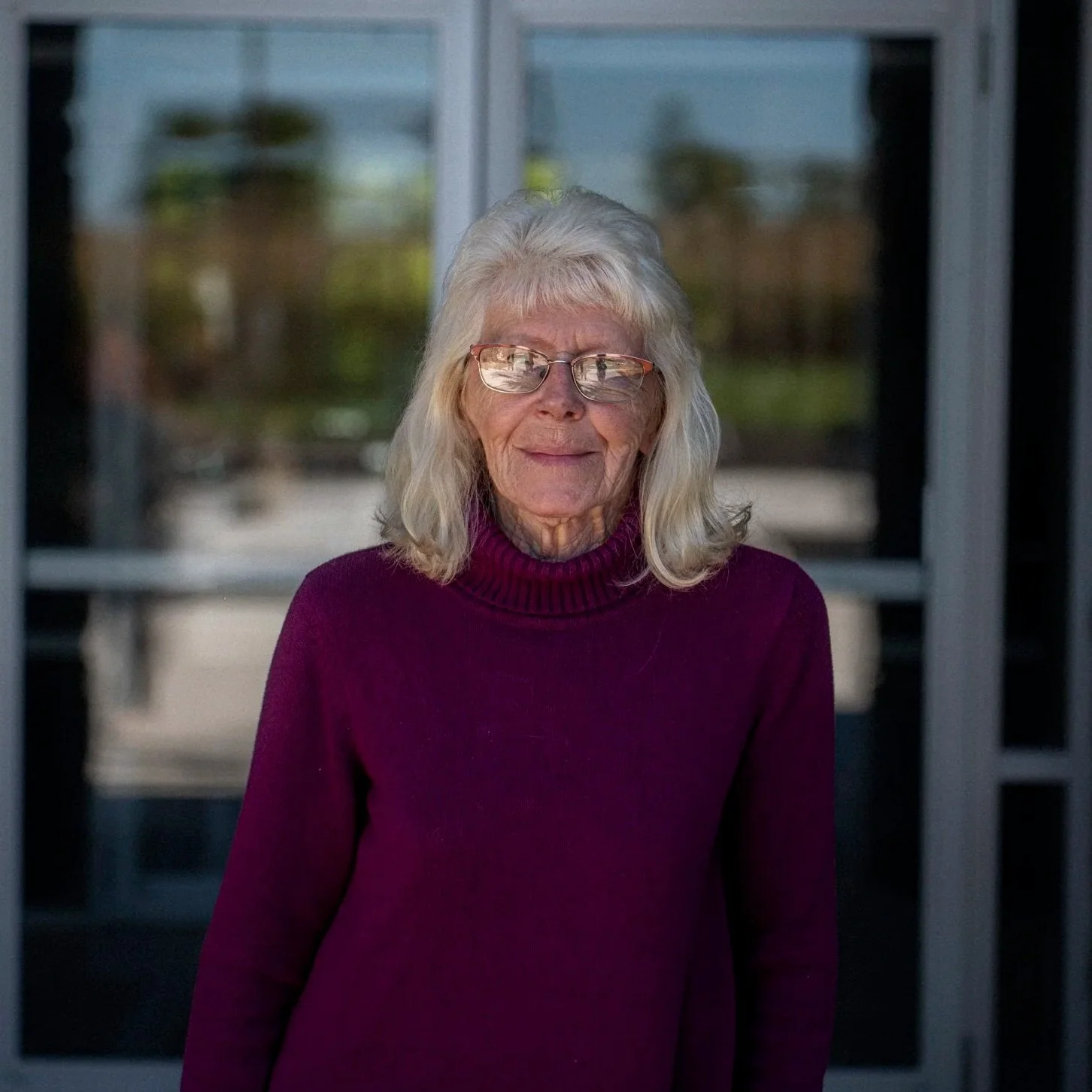 Portrait of an elderly woman with white hair, glasses, wearing a black sweater, standing against a light-colored brick wall.