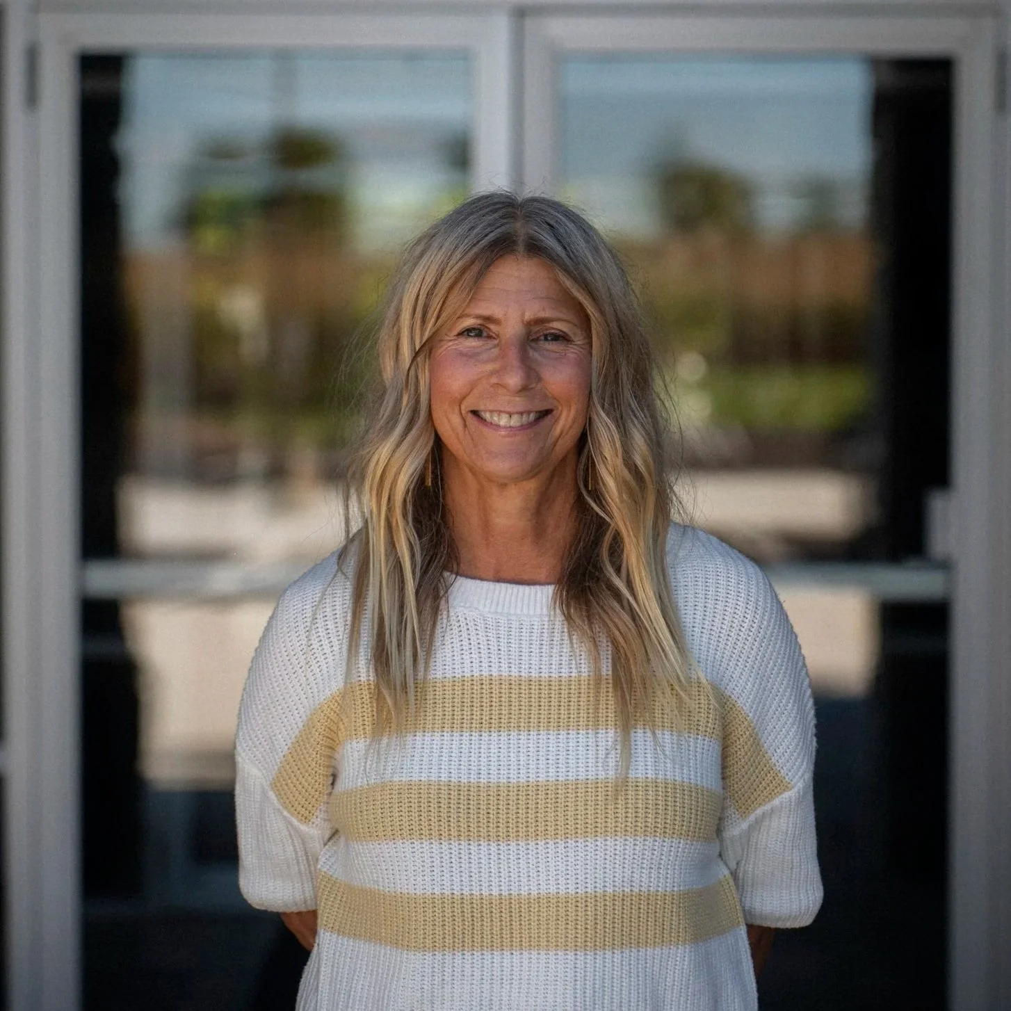 A woman with long blonde wavy hair, smiling, wearing a mustard yellow sweater, standing against a light gray brick wall.