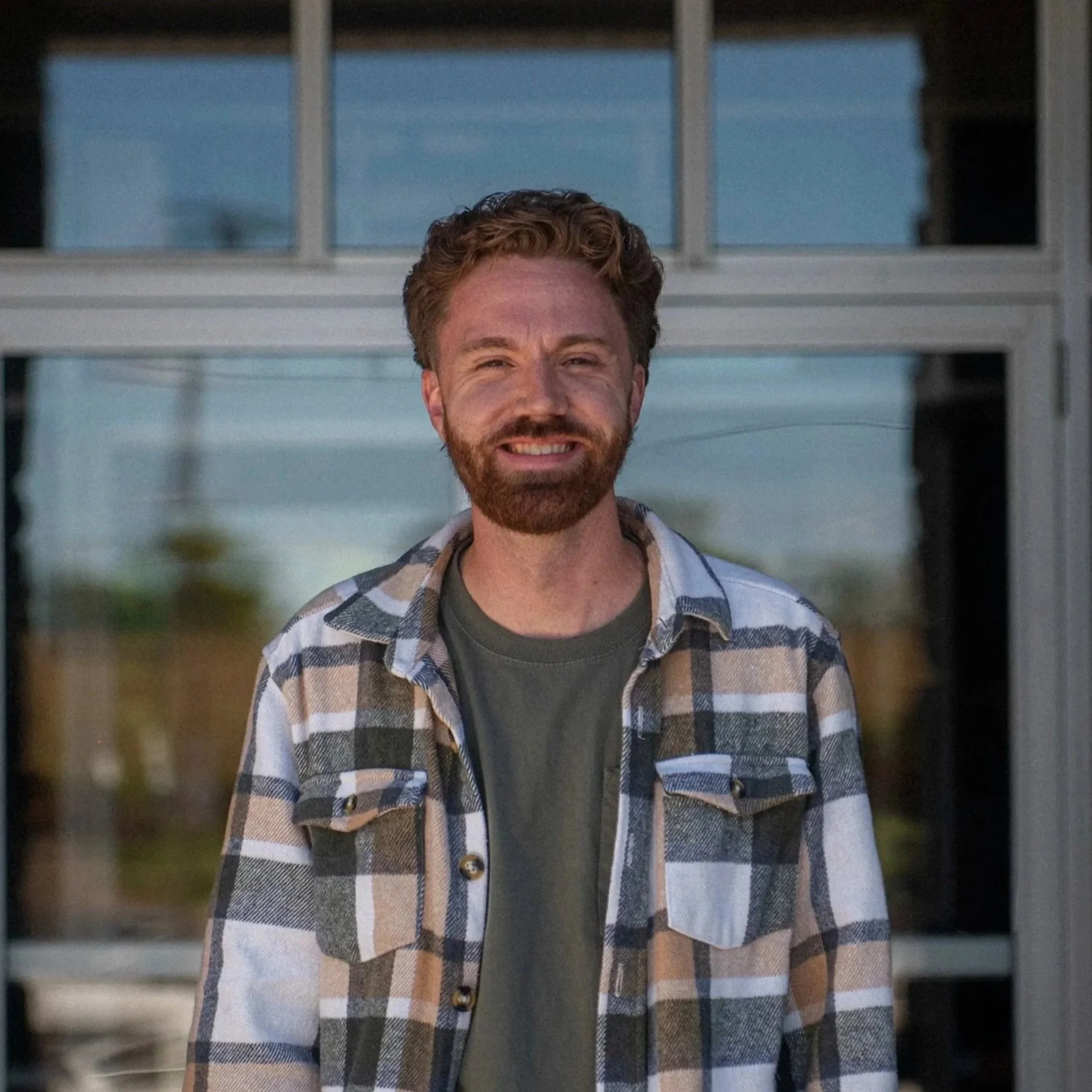 A man with a beard and wavy hair smiling, wearing a checkered jacket over a black shirt, standing against a light-colored background.
