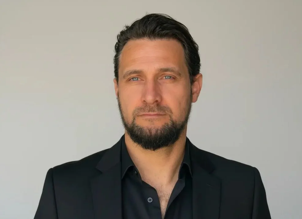 Ein Mann mit dunklem Haar, Bart, blaue Augen, trägt eine schwarze Anzugjacke und ein schwarzes Hemd, steht vor neutralem Hintergrund.