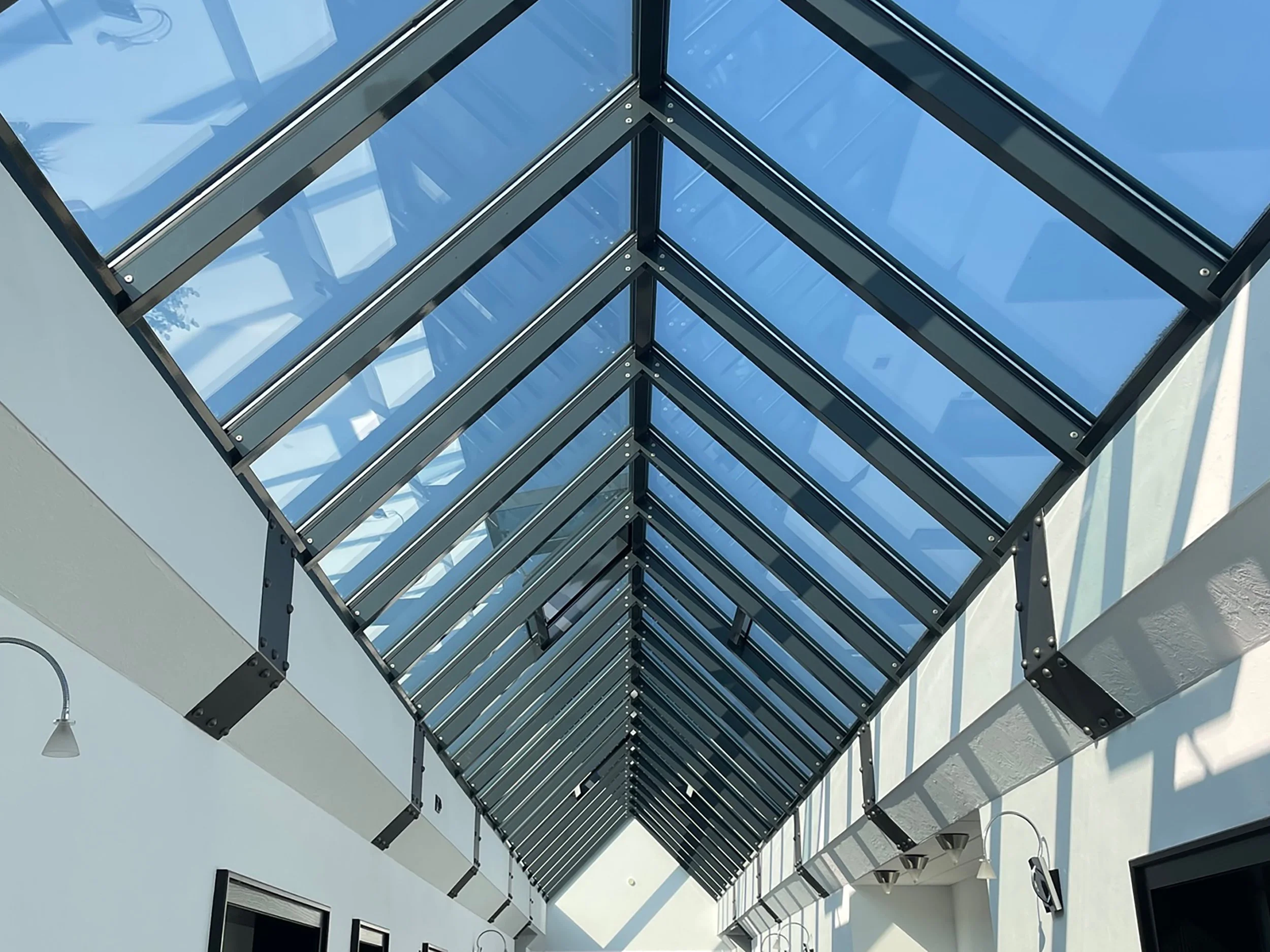 Blick nach oben auf ein Glasdach mit Metallskelett in einem Gebäude, durch das blaues Himmel zu sehen ist.