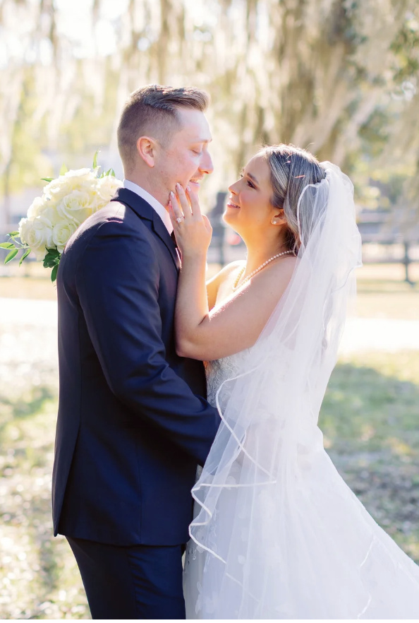 A bride and groom in wedding attire smiling and touching each other's faces outdoors.