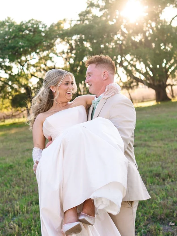 A bride and groom are outdoors on a sunny day, with the groom carrying the bride in his arms as they smile at each other. The background features trees and sunlight.