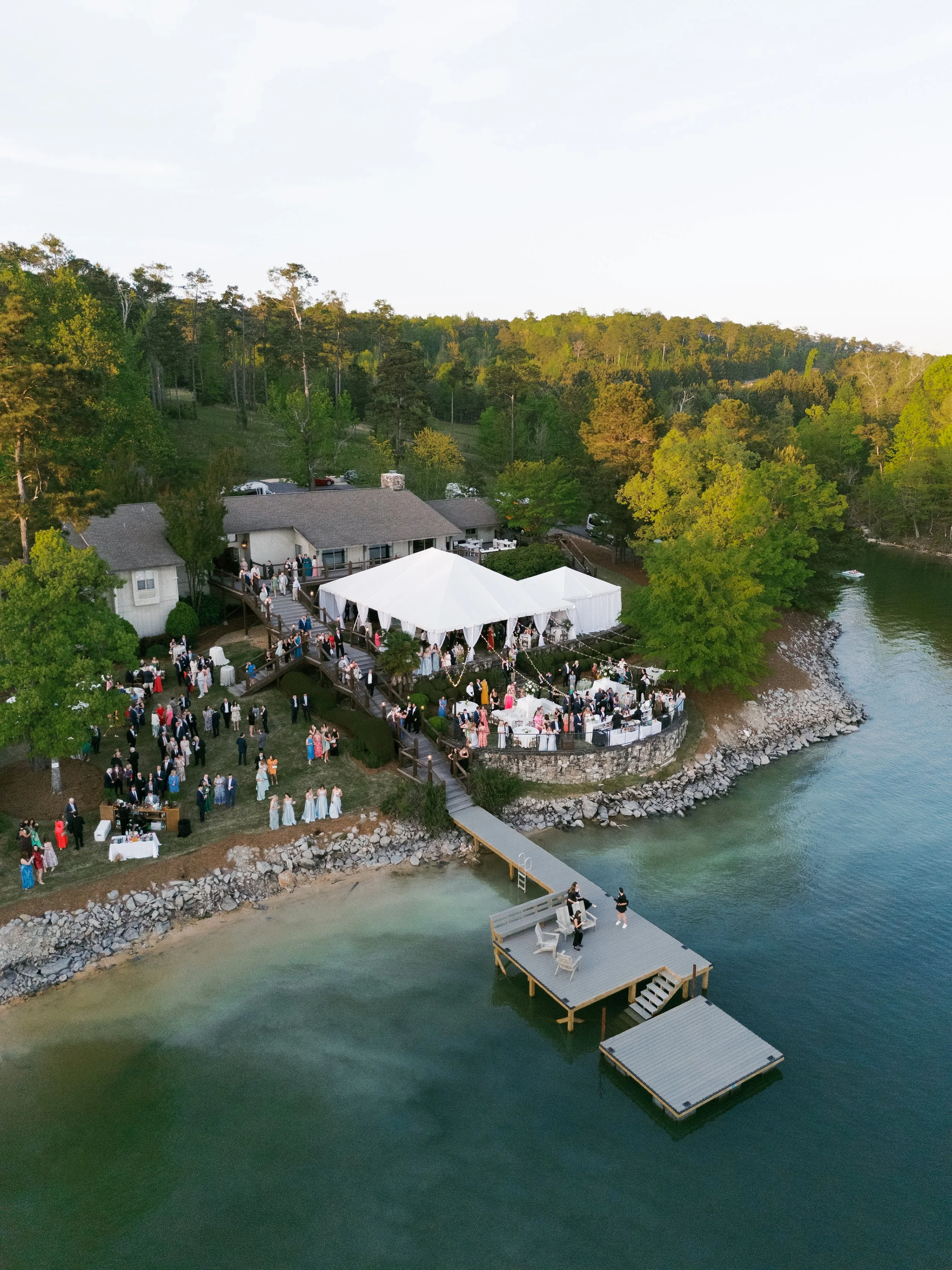 An outdoor wedding reception along a lakeshore, with guests on a deck, a white tent, and a wooden pier extending into the water surrounded by lush green trees.