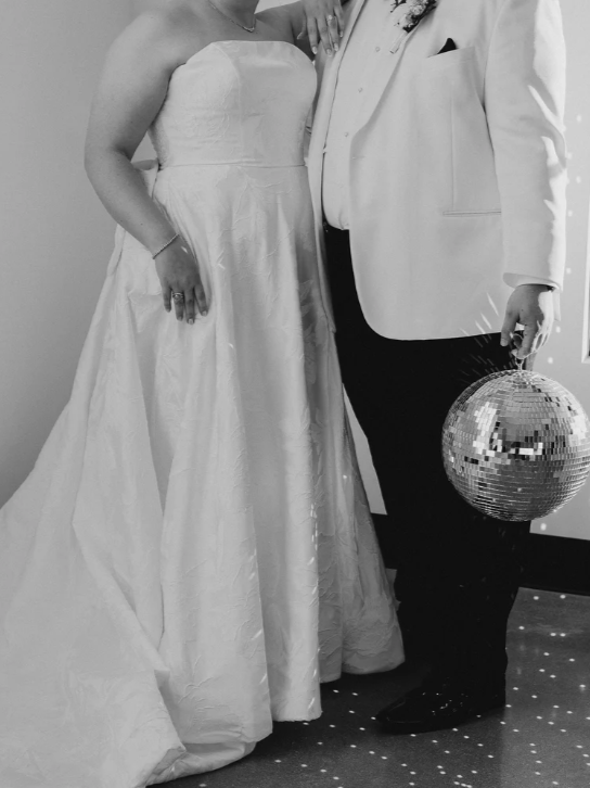 A bride and groom posing together at their wedding reception. The bride is wearing a strapless wedding gown, and the groom is dressed in a white tuxedo jacket with black pants, holding a disco ball.