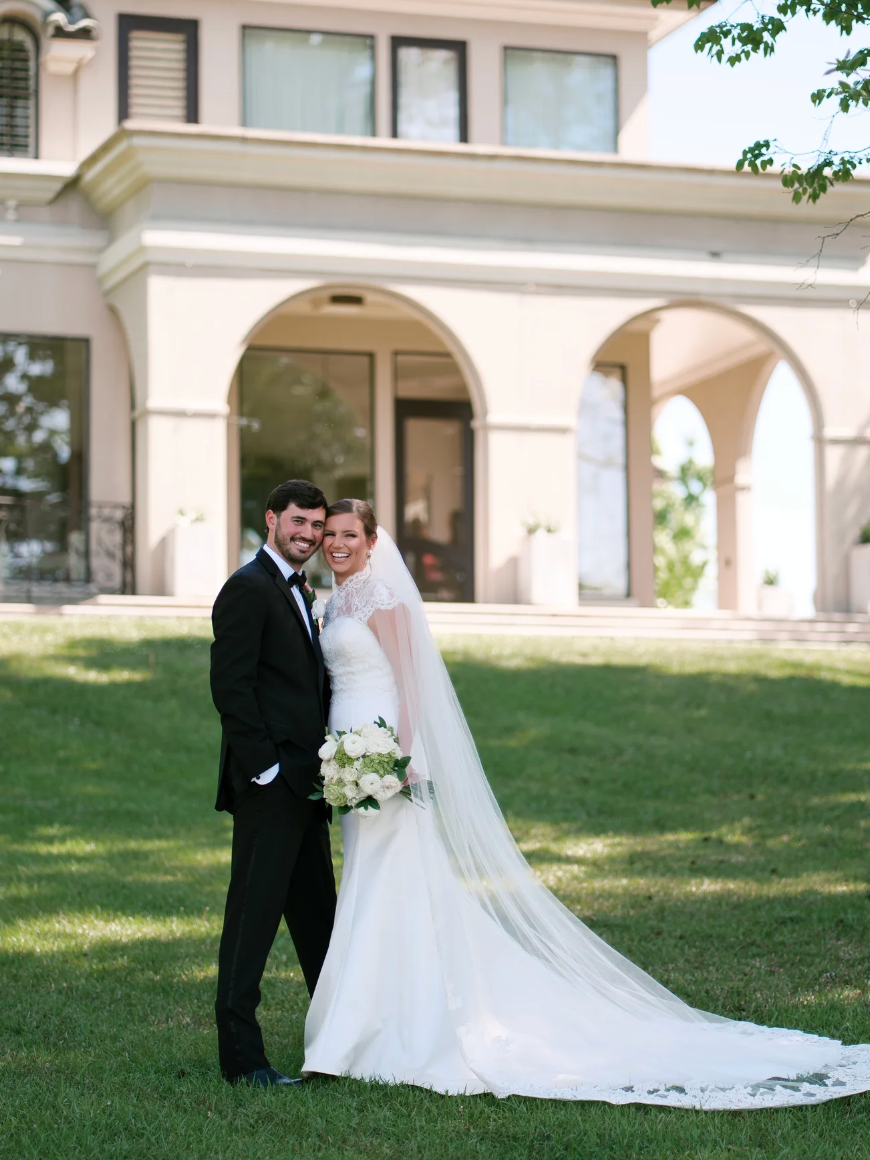 A bride and groom in wedding attire standing outdoors on a lawn in front of a large house with arched doorways. The bride is holding a bouquet of white flowers and wearing a white wedding gown with a long veil. The groom is dressed in a black tuxedo with a bow tie. Both are smiling and looking at the camera.