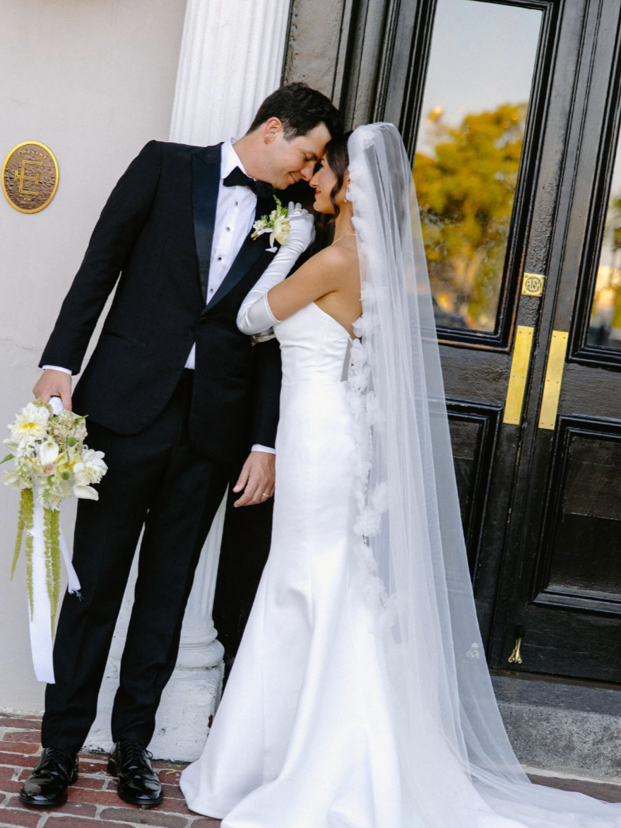 A bride and groom lean their foreheads together outside a building with black doors, dressed in wedding attire, with the bride wearing a white gown and veil, and the groom in a black tuxedo, holding a boutonniere and bouquet.