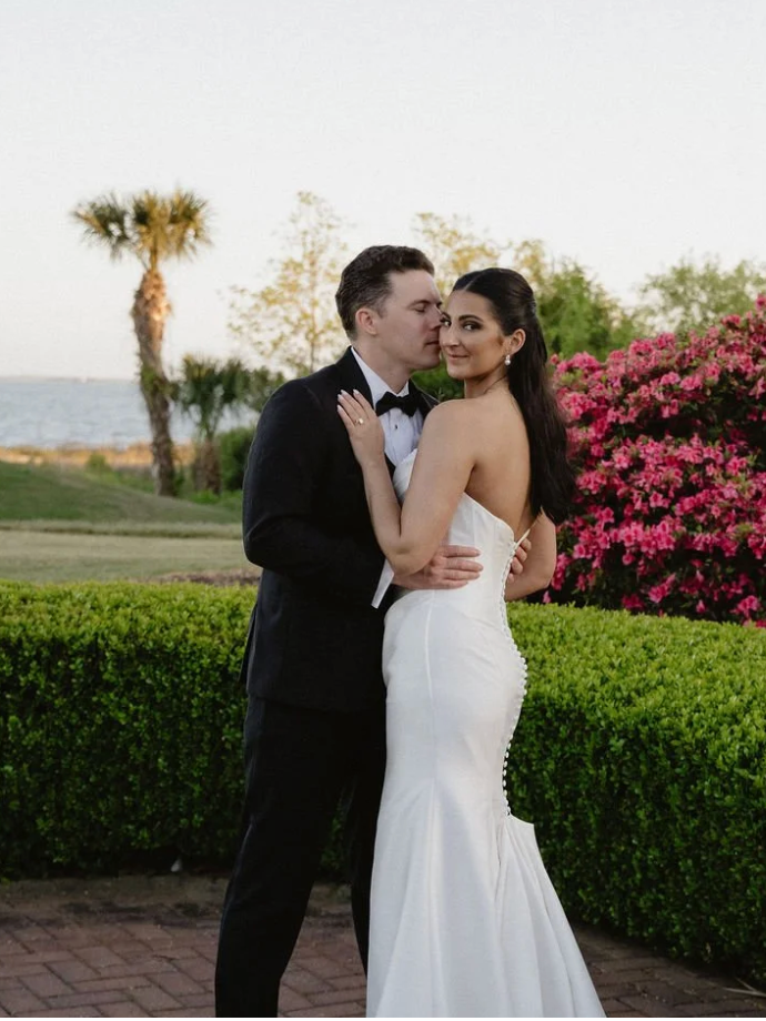 A newlywed couple, dressed in wedding attire, stands outdoors near a hedge and pink flowering bush, with water and palm trees in the background, during sunset.
