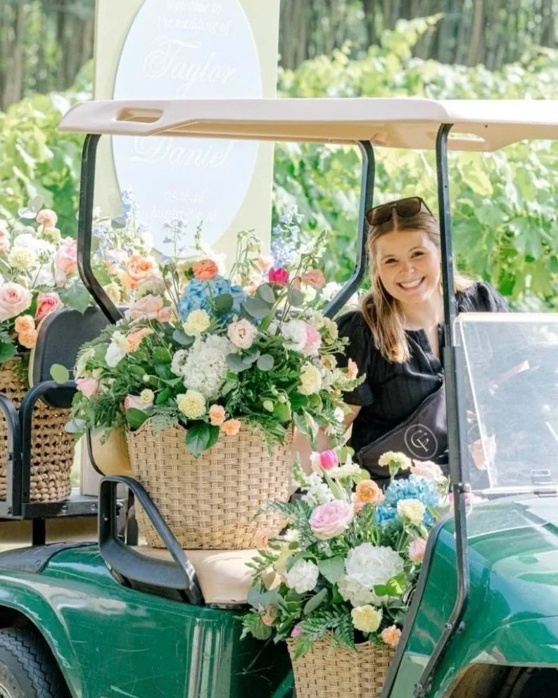 A woman sitting in a golf cart decorated with numerous colorful flowers, smiling outdoors in a garden setting.