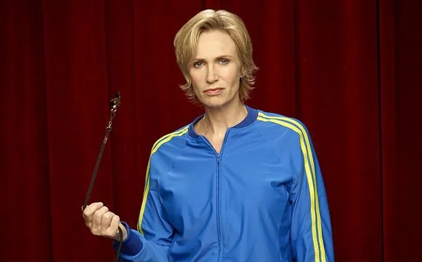 A woman with short blonde hair standing in front of a red curtain, wearing a blue sports jacket with yellow stripes and holding a baton.