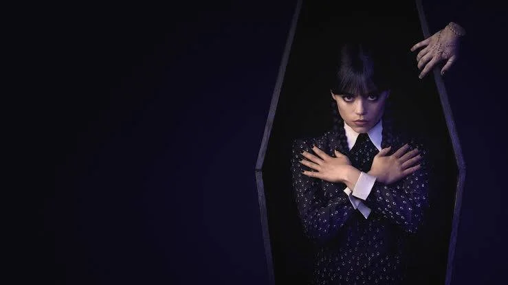 A woman with black hair and bangs peeking out of a coffin, crossing her arms. An older woman's hand is reaching in from above.