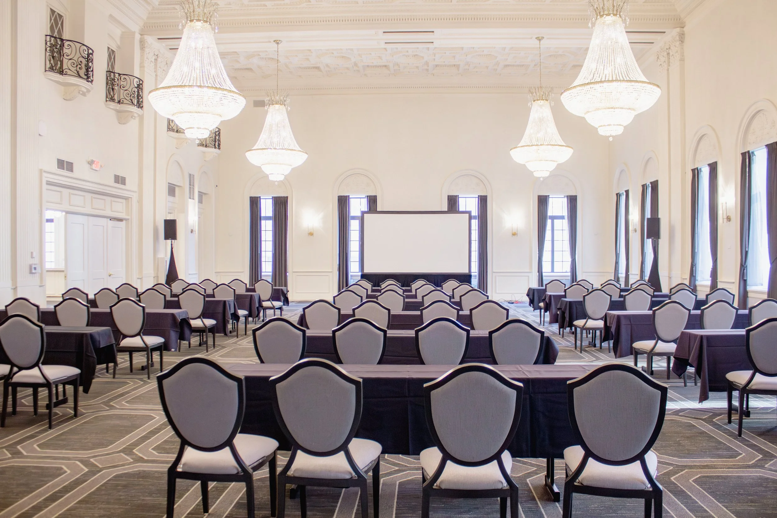 Large elegant conference room with high ceilings, multiple chandeliers, rows of chairs arranged facing a large projection screen, windows with curtains, and a patterned carpet.