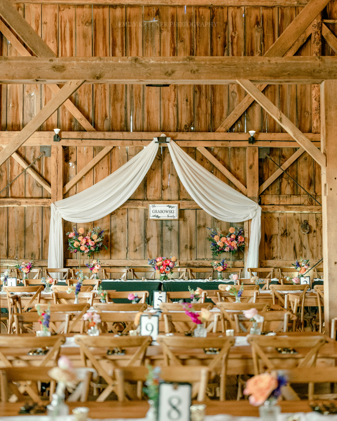 Indoor / outdoor rustic wedding reception with wooden tables and chairs decorated with small floral arrangements, a draped white fabric backdrop behind the head table,