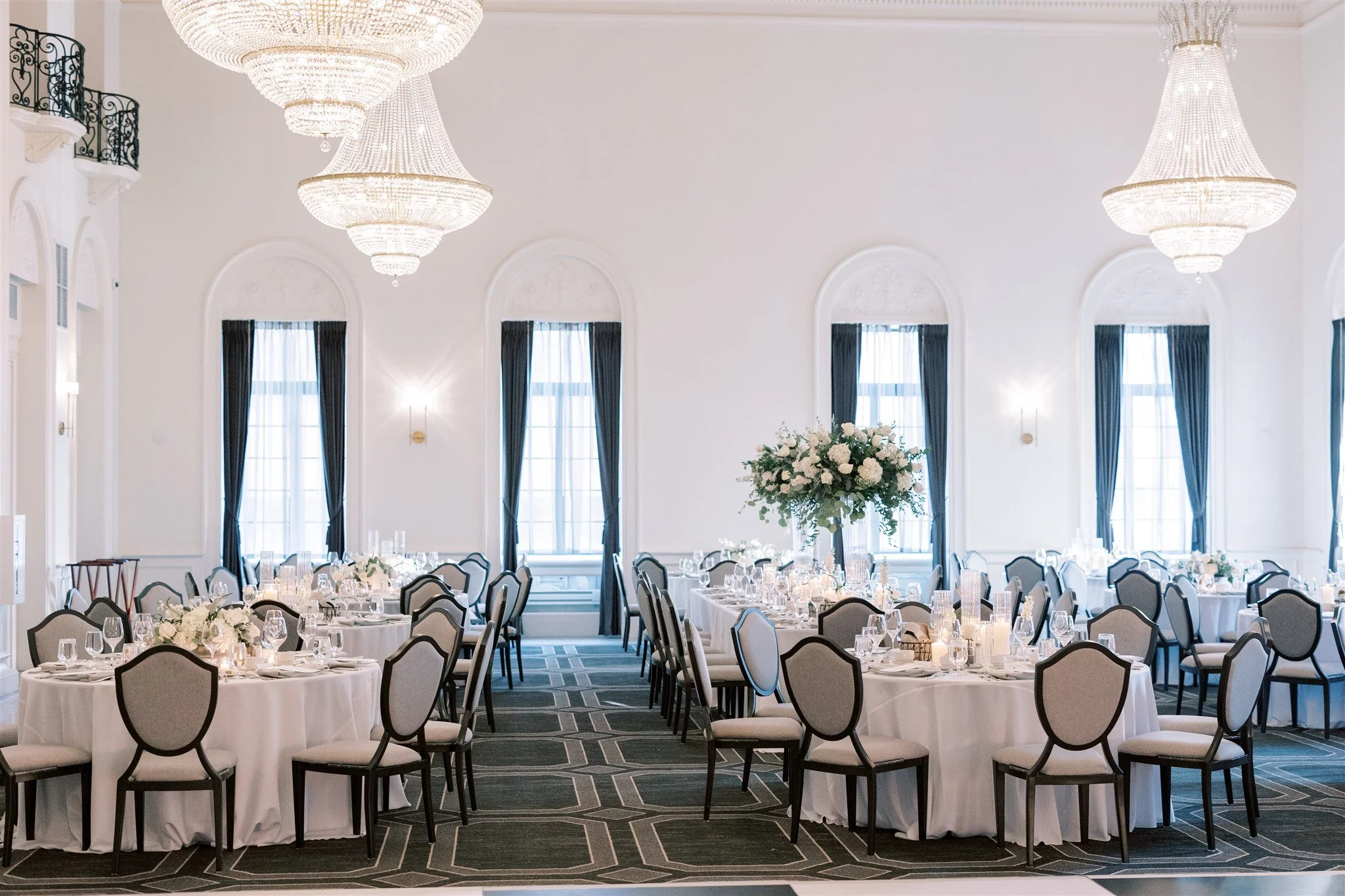 Elegantly set banquet hall with round tables covered with white tablecloths, floral centerpieces, and multiple glasses and place settings, illuminated by grand chandeliers and tall windows with dark curtains.