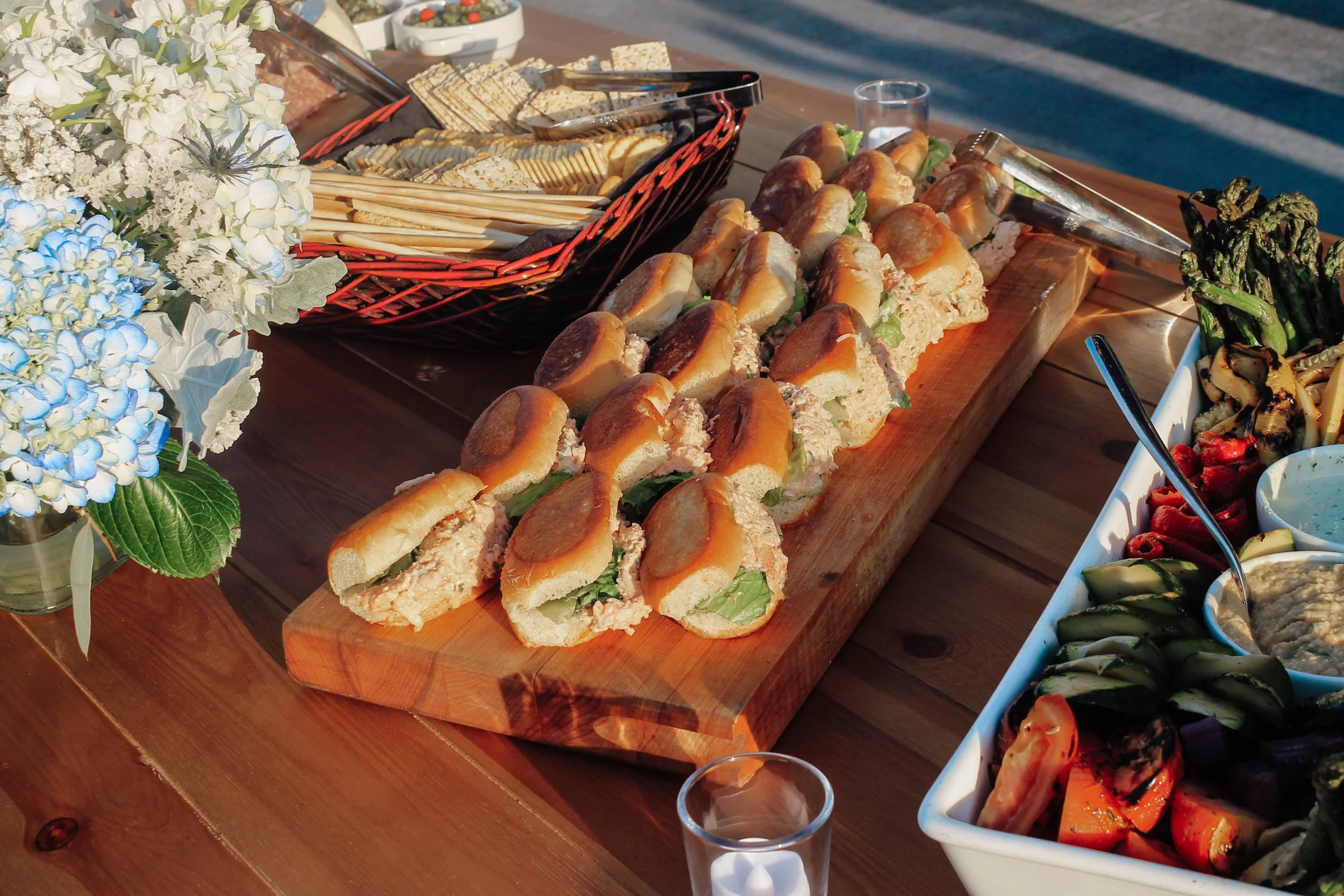 A picnic table with a platter of mini sandwiches filled with lettuce and chicken or tuna salad, next to a bowl of grilled vegetables including zucchini, red peppers, and eggplant, and a vase of white and blue hydrangea flowers in a glass jar.