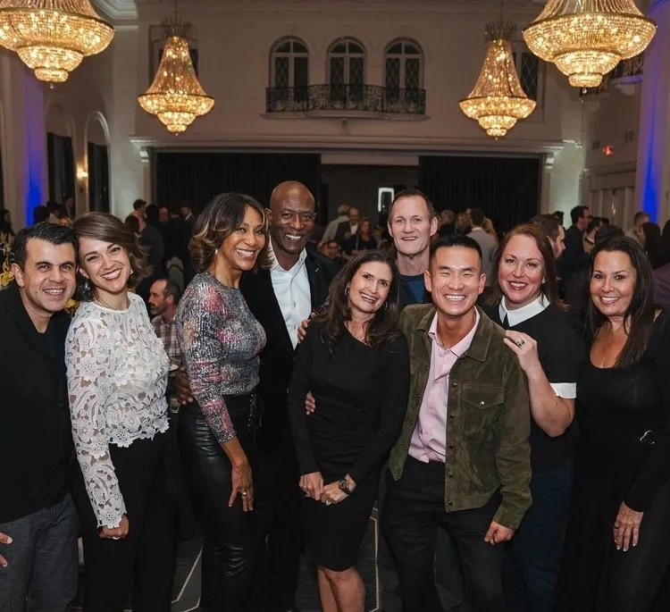 Group of people smiling and posing for a photo at a formal event in an elegant ballroom with chandeliers.
