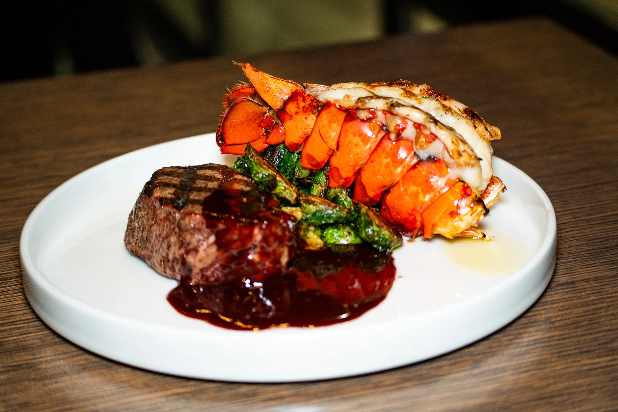 A surf and turf dish featuring grilled steak with sauce, lobster tail, and sautéed Brussels sprouts on a white plate.