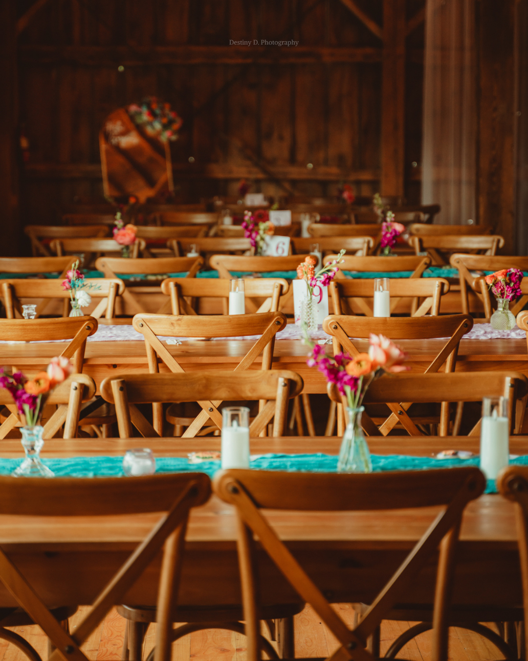 Wedding or event reception table setup in a rustic barn with wooden chairs, colorful flower centerpieces, candles, and table numbers.