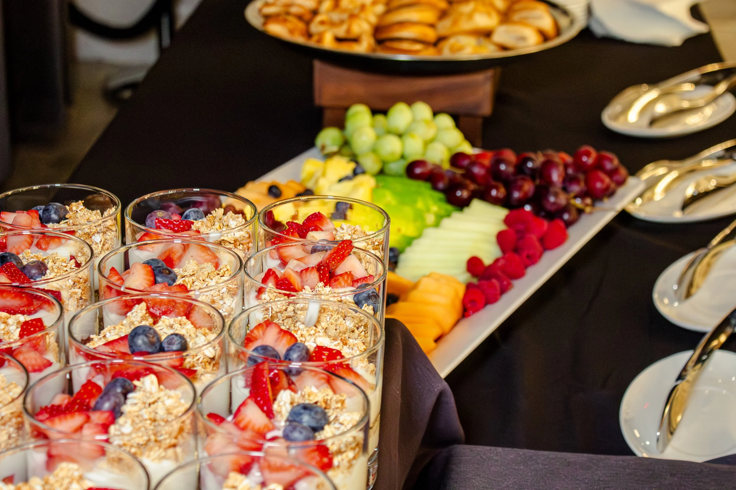 Close-up of parfait glasses filled with yogurt, topped with fresh strawberries, blueberries, and granola, arranged on a dark table at a buffet with assorted fresh fruits and snacks in the background.