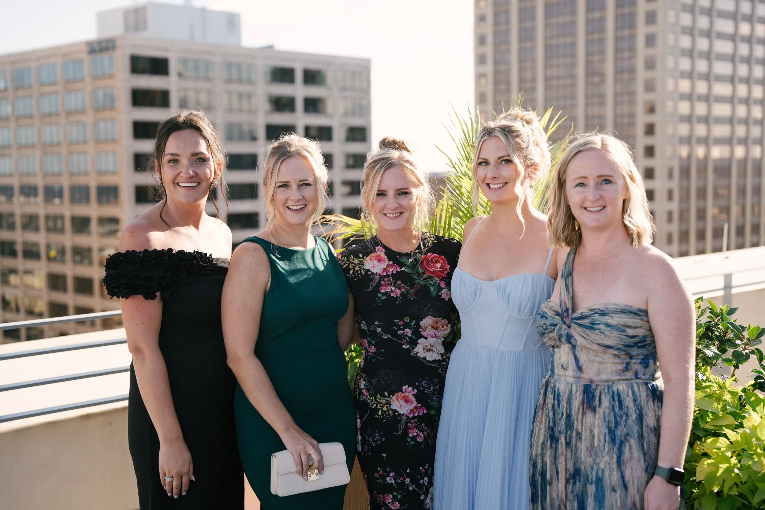 Group of five women standing on a balcony in front of a cityscape with tall buildings, dressed in formal attire, smiling at the camera.