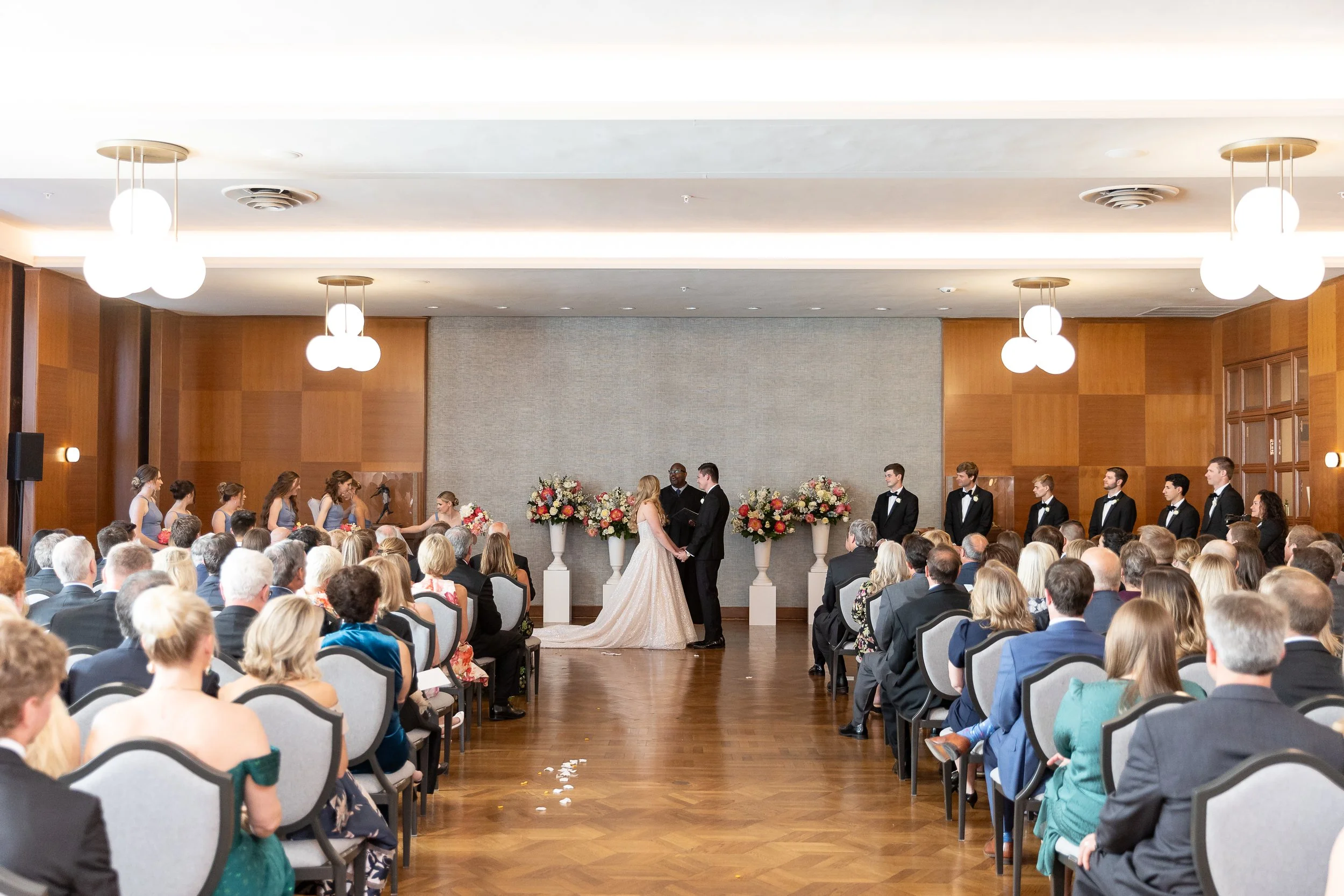 Wedding ceremony with a bride and groom exchanging vows in front of guests in an indoor venue with wood-paneled walls and hanging lights.