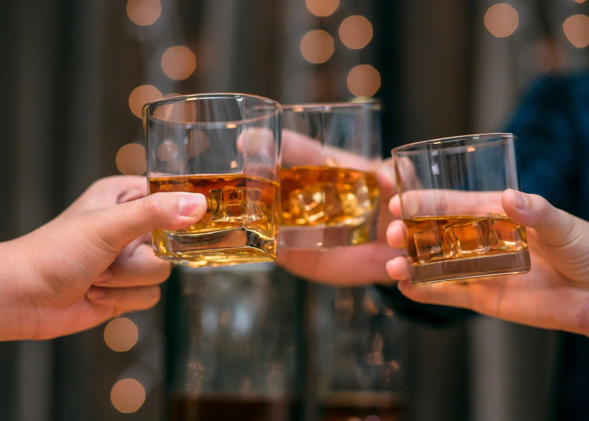 Three hands holding glasses filled with whiskey and ice, toasting with bokeh lights in the background.
