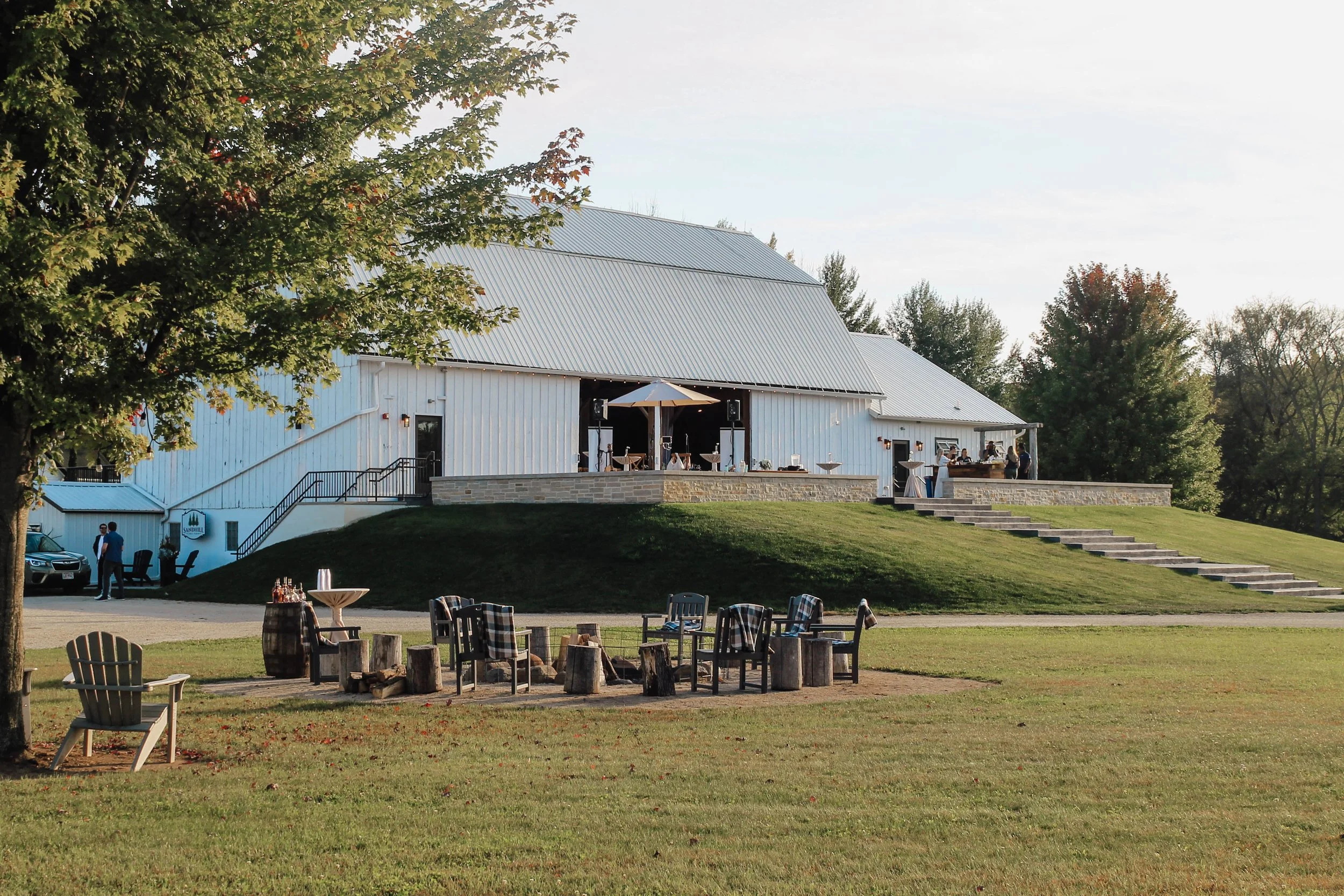White barn with metal roof and outdoor patio area set up with tables and chairs, surrounded by green grass and trees, likely during late afternoon.