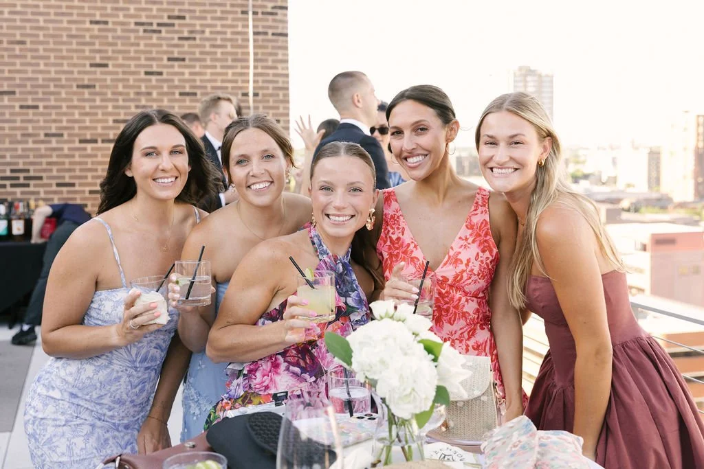 Five women smiling and holding drinks at a rooftop party with city skyline in the background.