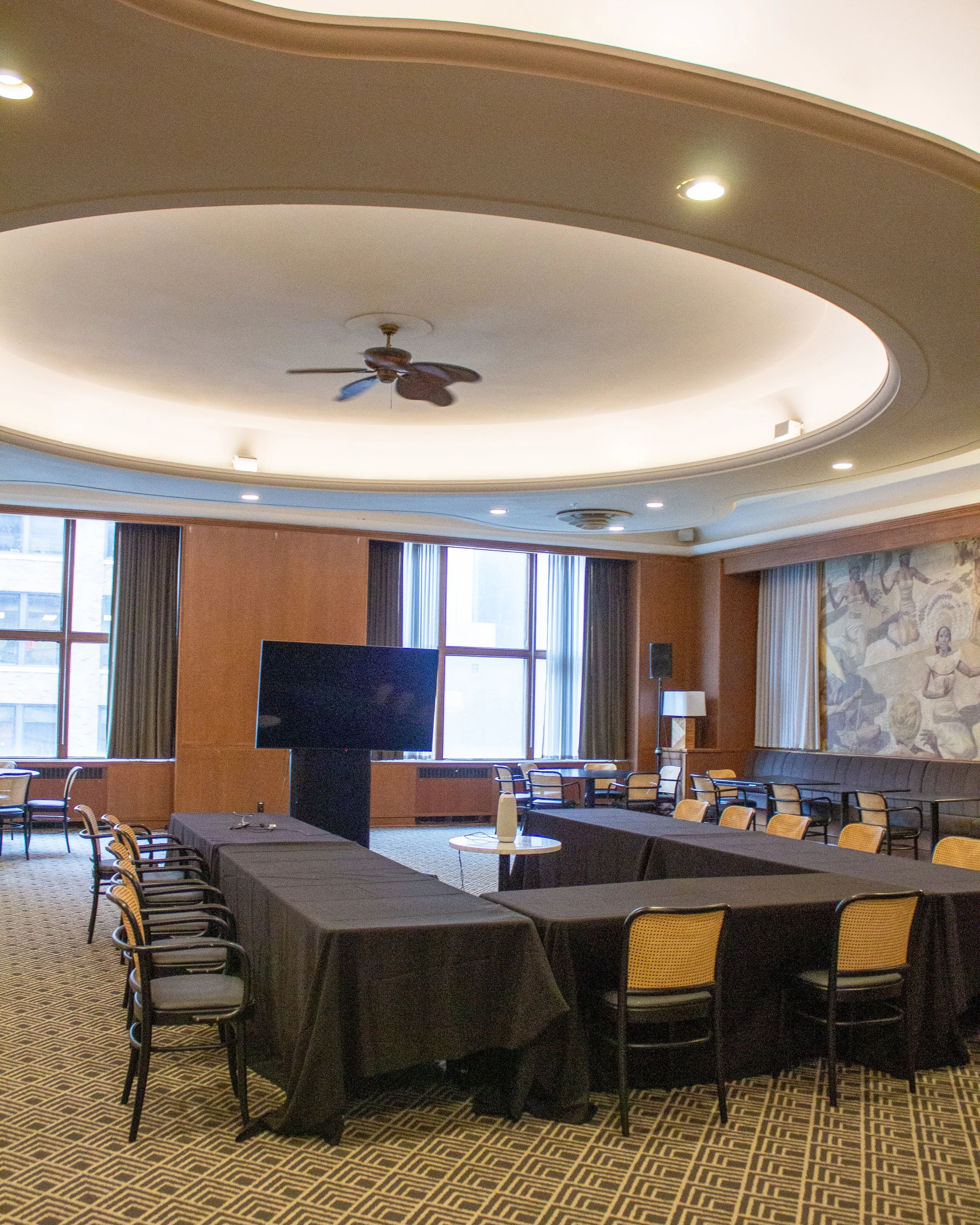 Conference room with round tables, black tablecloths, yellow chairs, large windows, patterned carpet, and a mural on the wall.