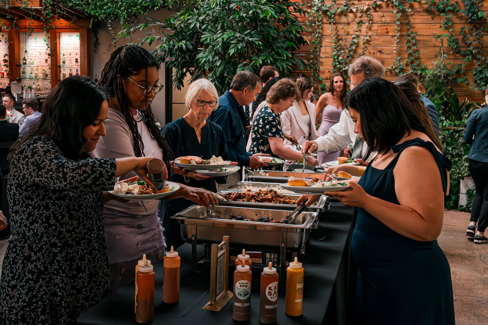 People serving themselves from a buffet at a social gathering, with various dishes and sauces available. The setting includes plants and a wooden backdrop, with attendees appearing to be engaged and enjoying the event.