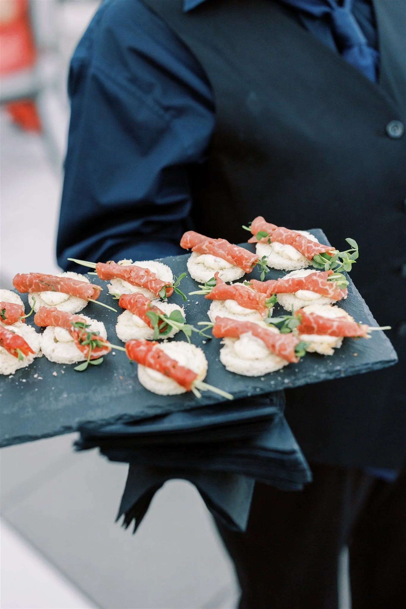 A waiter in a black uniform holds a tray with small appetizers topped with thin slices of cured meat, garnished with microgreens.
