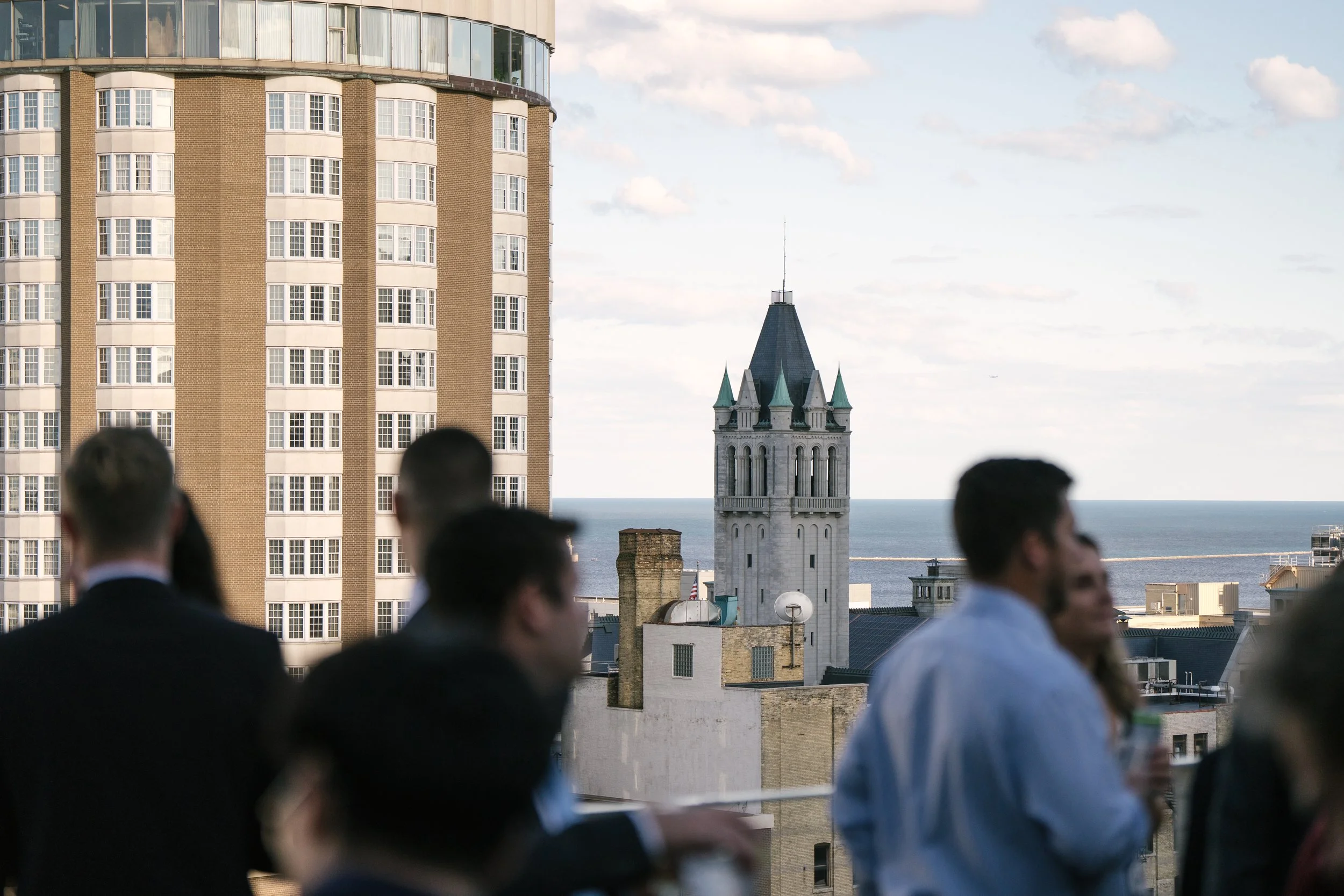 People are gathered on a rooftop or balcony, with a cityscape in the background, including a tall modern building and older church steeples, under a partly cloudy sky.